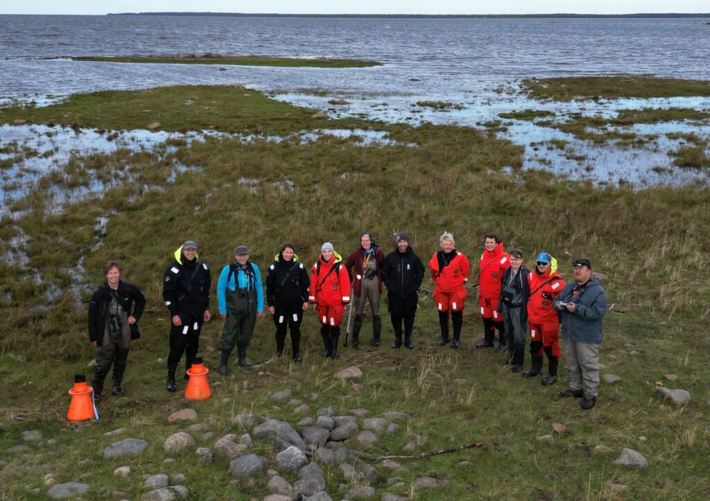 Group picture from above ground. Twelve people standing on a sea shore meadow.